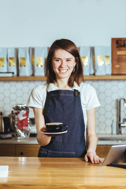 Amicale femme barista tenant une tasse de cappuccino dans un café. Concept de culture du café
. - Photo, image