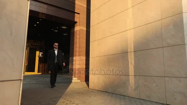 The fashionably dressed man in the dack suit, coat and sunglasses is keeping his hand in the pocket and leaving the building. - Video