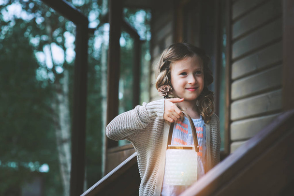child girl with candle holder relaxing in evening at cozy country house. Kid spending summer vacation in log cabin in the woods - Photo, Image