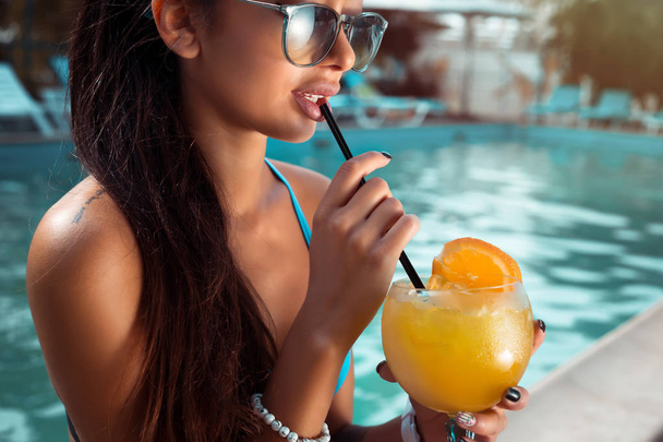 Beautiful brunette woman wearing a blue bikini and jeans shorts, enjoying the pool, summer time.Sensual brunette beautiful girl with perfect body drinking cocktail while relaxing near the pool - Photo, image