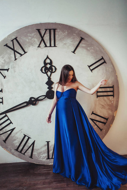 Waiting for child. Young brunette pregnant woman with long beautiful hair staying near the big clock in long blue dress, smiling and throws up her dress - Foto, imagen