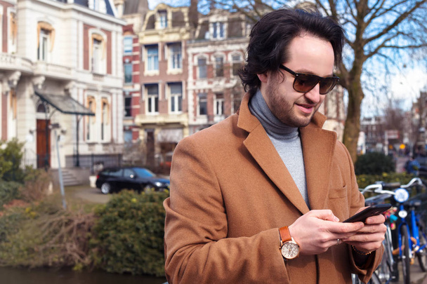 Moda model mężczyzna pozowanie w jesienny park. Pozowanie nadaje się człowiek. Młody biznesmen miejskich professional na smartfonie spaceru na ulicy za pomocą wiadomości sms SMS-y w aplikacji na smartfonie sobie kurtkę w Amsterdam - Zdjęcie, obraz