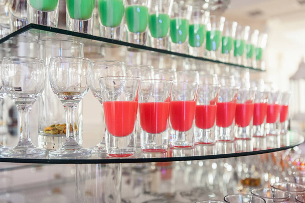 Ποικιλία από πολύχρωμες πράσινο και κόκκινο αλκοόλ βολές σε μικρά ποτήρια στέκεται στην γραμμή σ ' ένα γυάλινο περίπτερο - Φωτογραφία, εικόνα