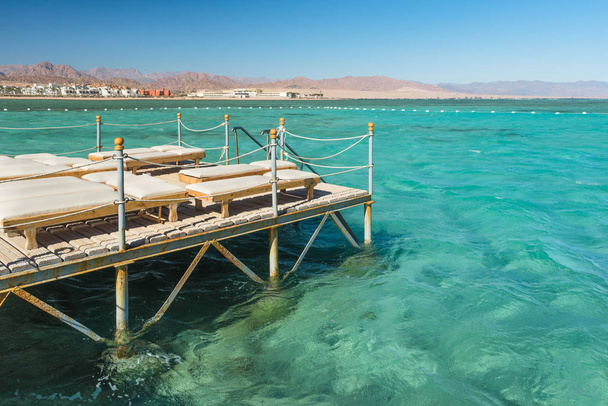 Pier with chaise longues in the sea in resort. Summer vacation. View at a clear sea with turquoise water. Summer vacation at a sea coastline in an exotic country. - Photo, Image