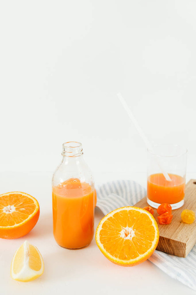 Láhev pomerančový fresh na bílém stole, vedle sklenice čerstvé a pomeranče s citronem. - Fotografie, Obrázek