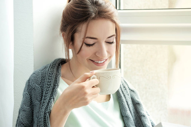 Bella giovane donna che beve tè vicino alla finestra a casa
 - Foto, immagini