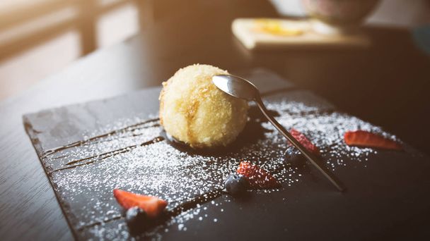 Grilled ice-cream in coconut with sauce and fruits on black stone plate with tea spoon. Asian food background. Eating concept. Restaraunt place with wooden table. Flare Copy space for text, design - Fotó, kép