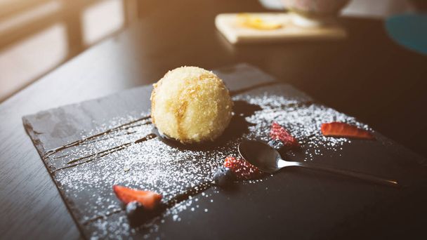 Grilled ice-cream in coconut with sauce and fruits on black stone plate with tea spoon. Asian food background. Eating concept. Restaraunt place with wooden table. Flare Copy space for text, design - Fotografie, Obrázek