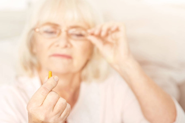 Thoughtful aging lady examining closely the pill at home - Valokuva, kuva