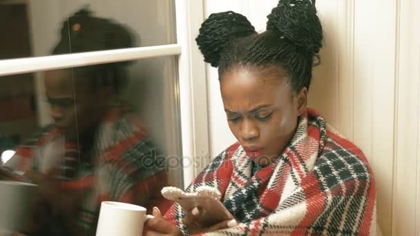 The horizontal close-up portrait of the surprised afro-american woman with african pigtails, reading articles via mobile phone and drinking tea under the red knitted plaid near the window. - Materiał filmowy, wideo