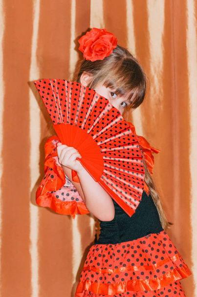 Cute little girl wearing beautiful red and black dress with matching headband, using hand fan, studio background - Foto, afbeelding