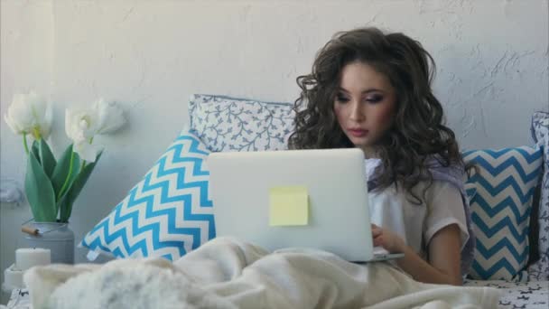 Νεαρή γυναίκα γράφει το κείμενο σε ένα φορητό υπολογιστή στην κρεβατοκάμαρα, που καλύπτεται με μια κουβέρτα. - Πλάνα, βίντεο