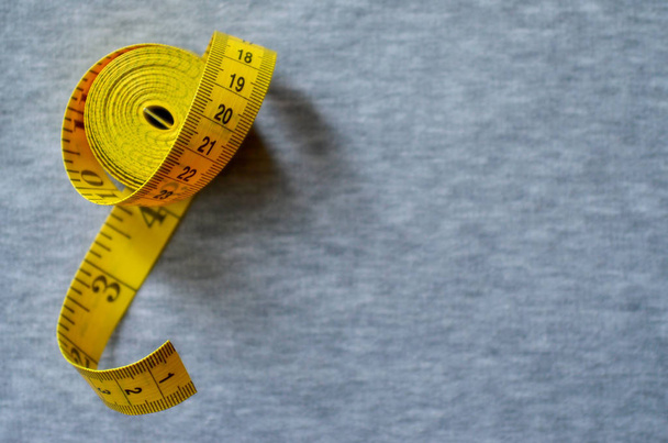 The measuring tape of yellow color with numerical indicators in the form of centimeters or inches lies on a gray knitted fabric. Background image about sewing sportswear for people, who slimming - Photo, Image