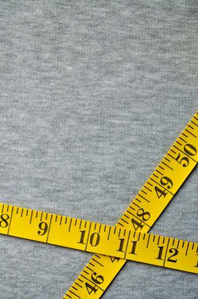 Η μεζούρα κίτρινο χρώμα με αριθμητικούς δείκτες με τη μορφή εκατοστά ή ίντσες βρίσκεται πάνω ένα γκρι πλεκτό ύφασμα. Φόντο έννοια για ράψιμο ρούχων για μεγέθη ειδικό φορέα - Φωτογραφία, εικόνα