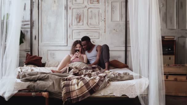 Multiracial couple in pajamas on the bed uses the smartphone. African man and european woman browse the Internet. - Imágenes, Vídeo