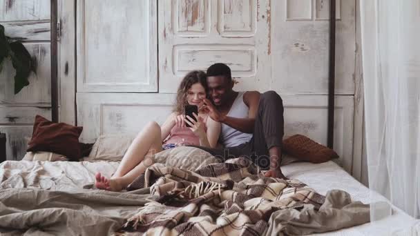 Multiracial couple in pajamas sitting on bed and use smartphone. African man and european woman looking photos, laughing - Footage, Video