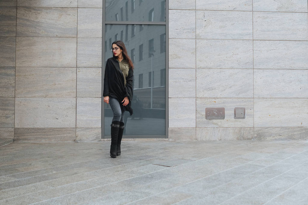 Fashion consept: beatiful young girl with long hair, glasses, red lips standing near modern wall wearing in green suit and grey jeans. She wanted talking on phone. - Foto, Imagem