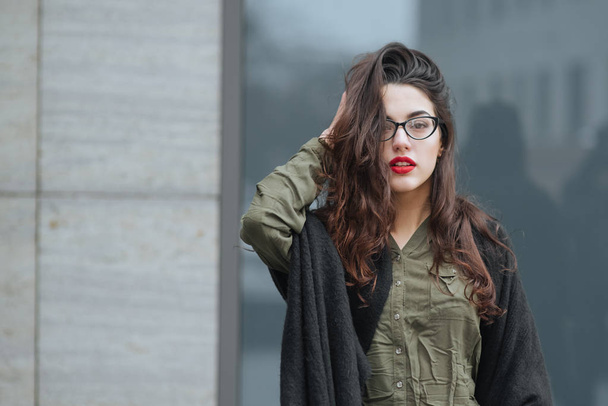 Fashion consept: beatiful young girl with long hair, glasses, red lips standing near modern wall wearing in green suit and grey jeans. She wanted talking on phone.  - Фото, зображення