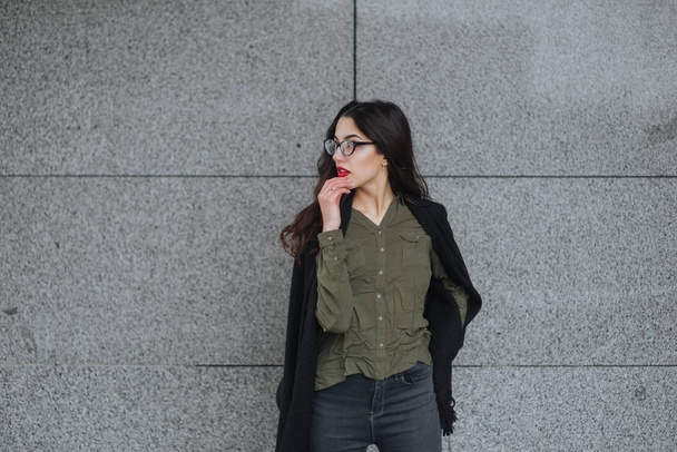 Fashion consept: beatiful young girl with long hair, glasses, red lips standing near modern wall wearing in green suit and grey jeans. She wanted talking on phone.  - Photo, image