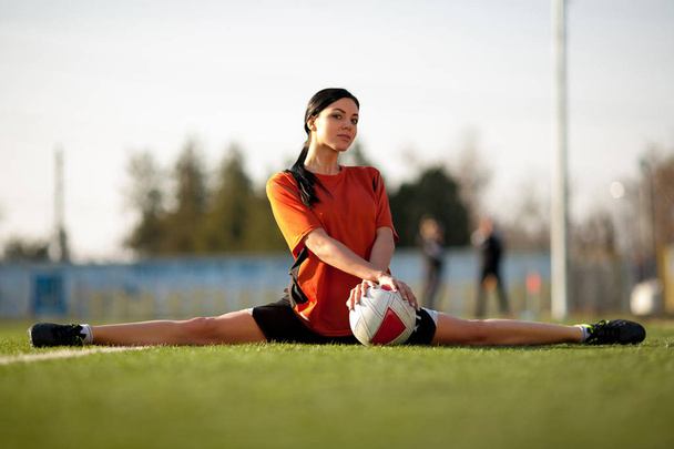 Дівчина у футбольній формі з м'ячем, сидить у гімнастичному шпагаті
 - Фото, зображення