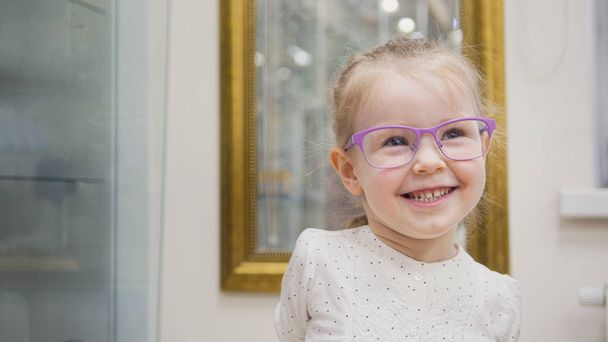 Niña rubia en gafas sonriendo y juega en la clínica de oftalmología
 - Foto, Imagen