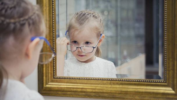 Маленька дівчинка пробує нові окуляри біля дзеркала - покупки в офтальмологічній клініці
 - Фото, зображення