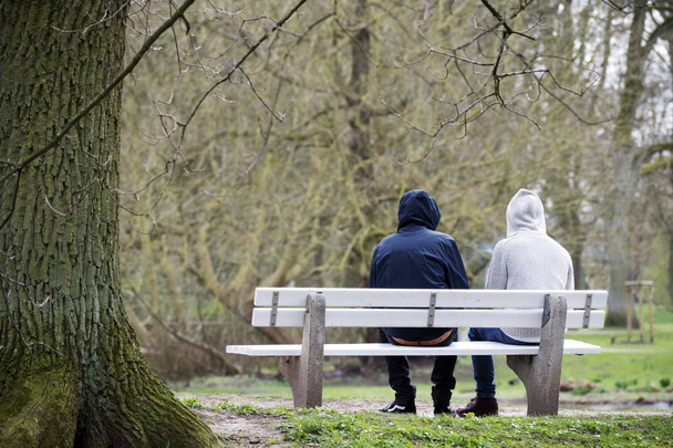 Two young men with hoods sitting together on a bench from behind in bad weather in the park - Foto, imagen