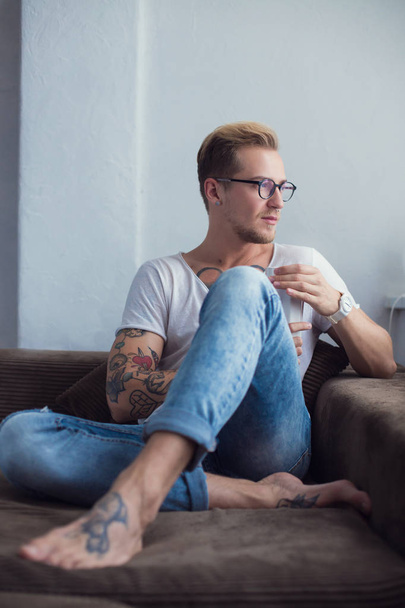 Young fashionable model sitting on sofa at home and drinking cacao. Relax. Tattoo - Foto, Bild
