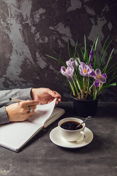 The girl's hands closeup with a pen in hand writing in a notebook. On the table is a Cup of coffee and a vase with flowers crocuses - Zdjęcie, obraz