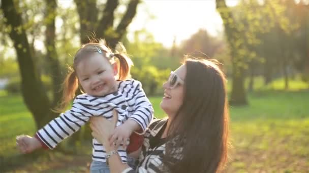 Openlucht portret van gelukkige moeder en dochter in het Park in het voorjaar. Mooie Brunette met zonnebril is uitvoering schattig meisje op handen en haar wervelende. - Video