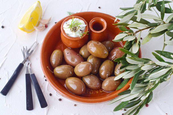 Olives in a traditional brown ceramic bowl, with tzatziki sauce, lemon, garlic, olive branch and olive oil on a white abstract background - Foto, Bild