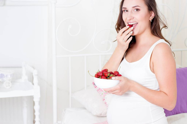 glückliche schwangere Frau auf dem Bett sitzend. Eine junge schwangere Frau sitzt im Schlafzimmer. Schwangere entspannt. junge schwangere Frau isst Erdbeeren. - Foto, Bild