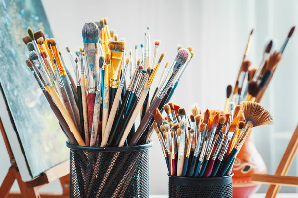 Artistic equipment in studio: wooden easel,  paintbrushes, tubes of paint, palette and paintings. - 写真・画像