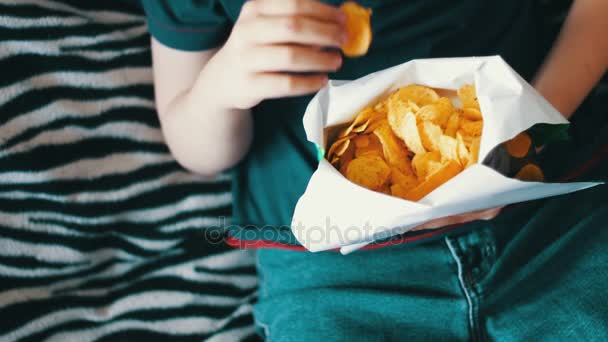 Niño adolescente comiendo papas fritas con las manos en el sofá en casa
 - Metraje, vídeo
