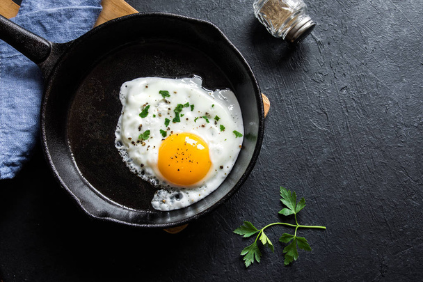 Fried egg. Close up view of the fried egg on a frying pan. Salted and spiced fried egg with parsley on cast iron pan and black background. - Fotó, kép