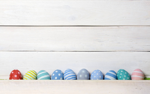 Десять разноцветных пасхальных яиц ручной работы выстроились в ряд на белом деревянном фоне. Плоский стиль
 - Фото, изображение