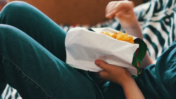 Έφηβος αγόρι τρώει τσιπς πατάτας με τα χέρια στον καναπέ στο σπίτι - Πλάνα, βίντεο