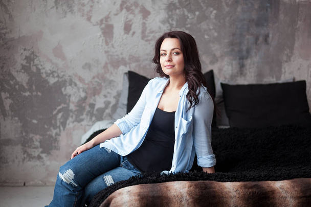 Sensual woman with dark hair in blue shirt and jeans sitting on a bed at home. Loft style interior - Fotografie, Obrázek