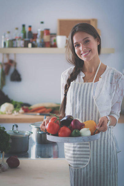 Souriante jeune femme tenant des légumes debout dans la cuisine - Photo, image