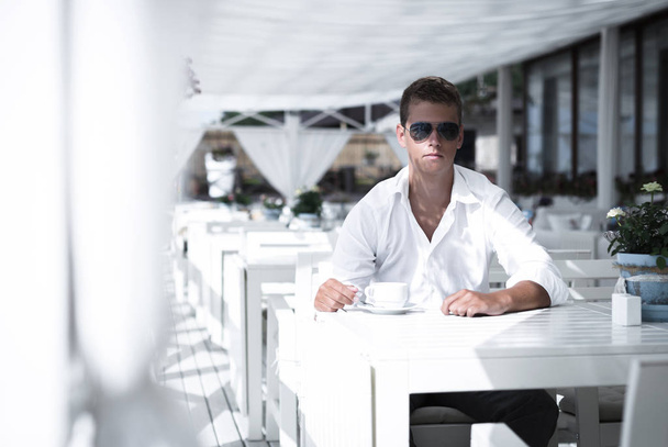 Young serious business style male is drinking coffee in seaside restaurant. Sitting alone wearing stylish sunglasses and white shirt Caucasian guy is looking forward at the camera. - Foto, imagen