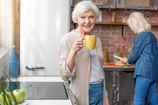 Cheerful old lady enjoying hot beverage in kitchen - Фото, изображение