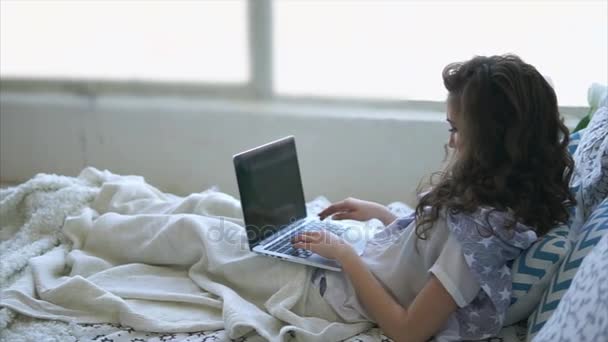 Adult girl prints text on laptop keyboard, lying under a blanket in bed. - Footage, Video