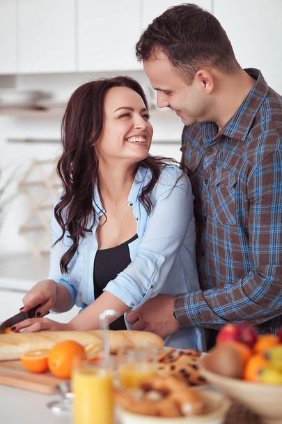 Couple making fresh organic juice in kitchen together. A young woman in a blue shirt slices a baguette. A man is hugging his girlfriend in a white kitchen. Romantic concept - Foto, afbeelding
