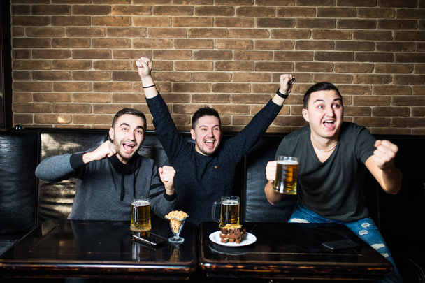 Νεαροί άνδρες πίνουν μπίρα, τρώνε σνακς και επευφημίες για αγώνα ποδοσφαίρου. Κερδίζοντας συναισθήματα. - Φωτογραφία, εικόνα