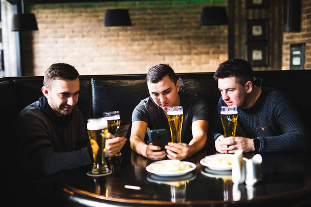Χαρούμενα παλιούς φίλους, διασκεδάζοντας με smartphone και πίνοντας μπύρα σε παμπ. - Φωτογραφία, εικόνα