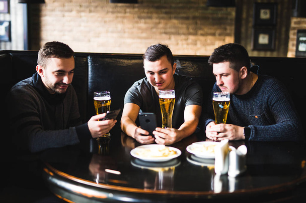 Χαρούμενα παλιούς φίλους, διασκεδάζοντας με smartphone και πίνοντας μπύρα σε παμπ. - Φωτογραφία, εικόνα