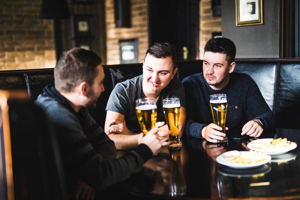 Trois jeunes hommes en vêtements décontractés parlent, mangent des chips et boivent de la bière pendant qu'ils sont assis dans un pub et discutent
. - Photo, image
