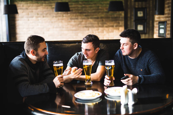 Τρεις νεαροί άνδρες στη casual ρούχα μιλάμε, τρώγοντας τσιπς και πίνοντας μπύρα, ενώ κάθεται σε παμπ και συζητώντας. - Φωτογραφία, εικόνα