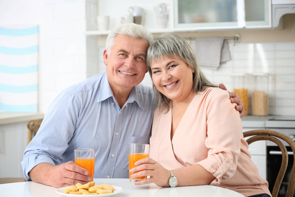 Beautiful mature couple with orange juice sitting at kitchen table - Foto, Imagem