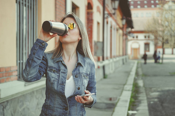 Attractive blonde young woman walking on the street, using her smartphone and drinking coffee - Foto, imagen